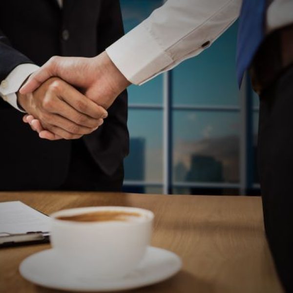 Two professionals shaking hands across a desk with cityscape in the background.